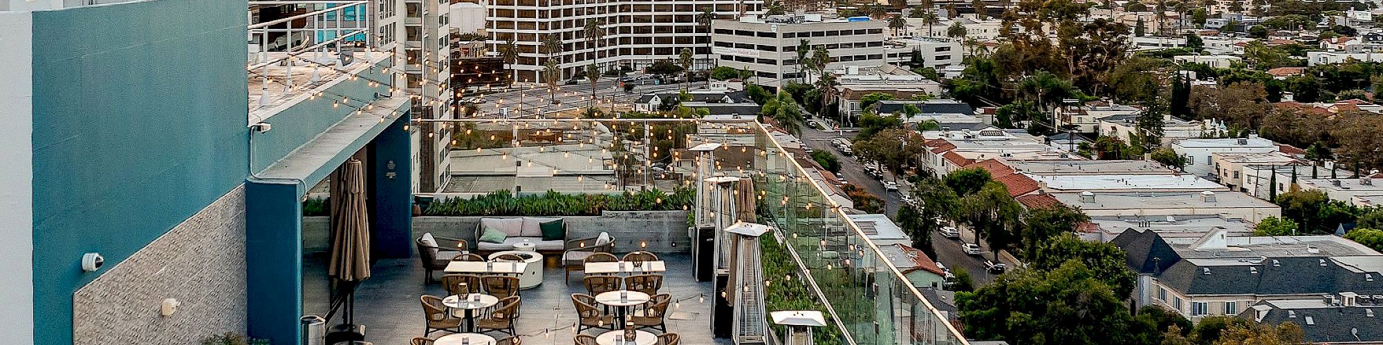 The image shows a rooftop terrace with tables and chairs overlooking a cityscape, surrounded by modern buildings and a vast skyline.