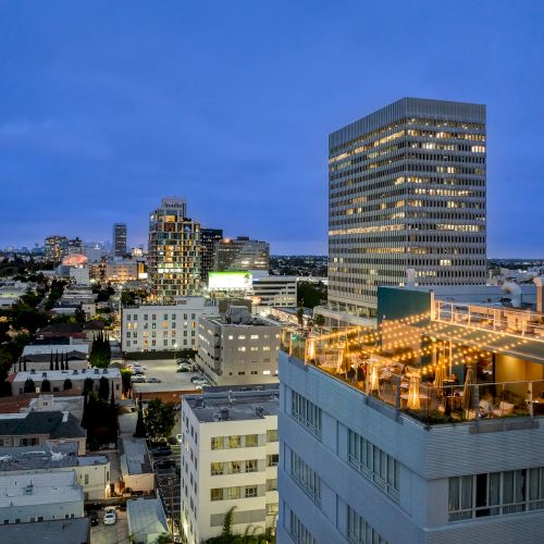A cityscape at dusk featuring buildings, with a lit rooftop area on the right side adding warmth to the scene.
