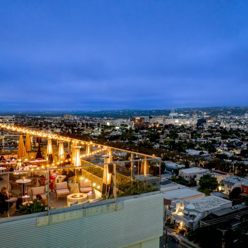 A rooftop bar with warm lighting overlooks a cityscape at dusk, featuring tables, umbrellas, and patrons enjoying the view.
