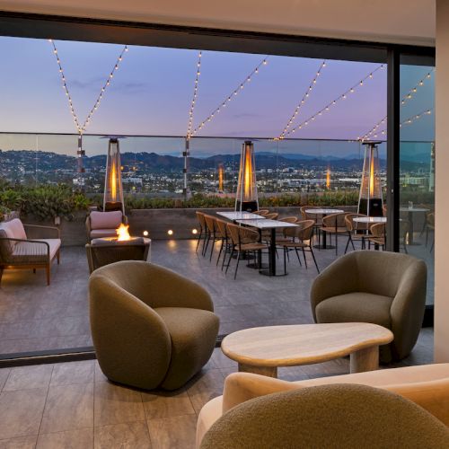 The image shows a modern indoor seating area with chairs and tables, opening to an outdoor terrace decorated with lights and heaters.