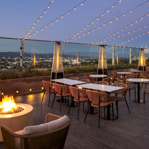 A rooftop patio with string lights, tables, chairs, and a fire pit, offering a view of the cityscape at dusk.