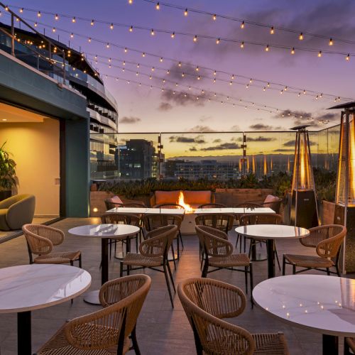 A rooftop patio at sunset with string lights, tables, wicker chairs, and outdoor heaters, creating a cozy and inviting atmosphere.