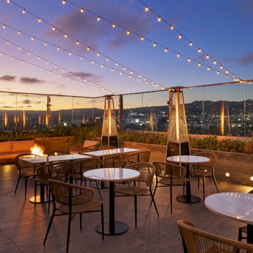 A rooftop patio with tables, chairs, string lights, and heaters overlooks a cityscape at sunset.
