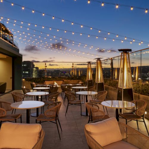 A rooftop patio at sunset with string lights, tables, chairs, and outdoor heaters, offering a cityscape view.