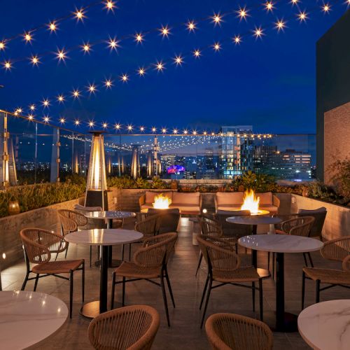 Rooftop patio with tables, chairs, and strings of lights at night; heaters and cityscape provide ambiance.
