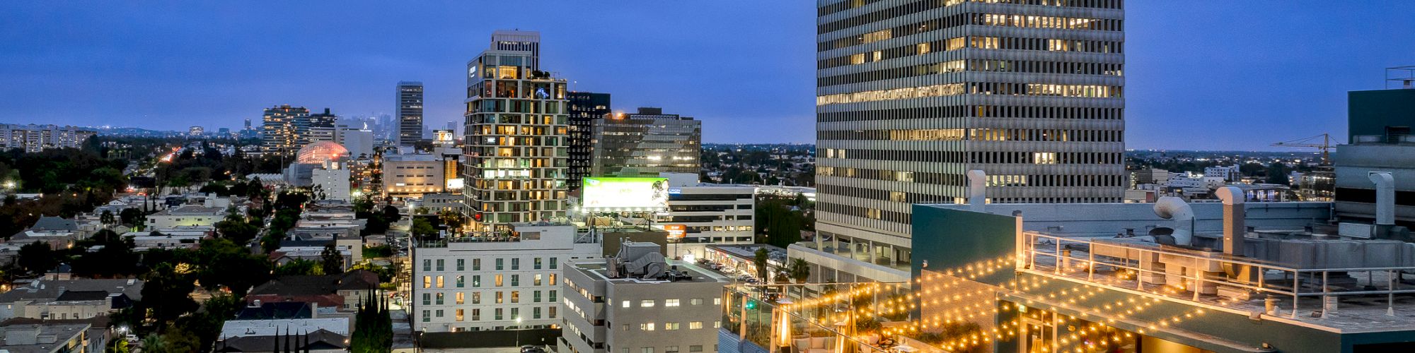 A cityscape at dusk with buildings, lit windows, and a rooftop decorated with string lights.