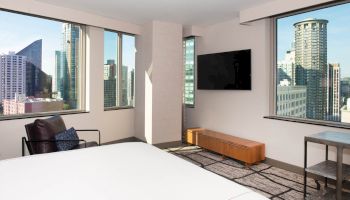 The image shows a modern hotel room with a large bed, TV, and city views through multiple windows. A chair and table are also visible.