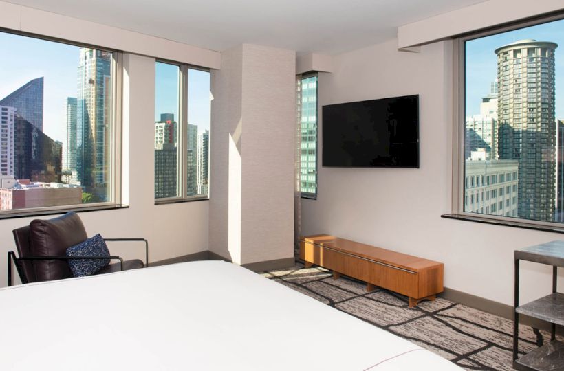 The image shows a modern hotel room with a large bed, TV, and city views through multiple windows. A chair and table are also visible.
