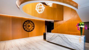 The image shows a modern lobby with a marble reception desk, wooden wall, circular clock, spiral staircase, and decorative lighting fixture.