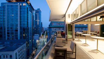 A modern rooftop terrace at dusk with seating and tables, overlooking a cityscape with illuminated skyscrapers and a street below.