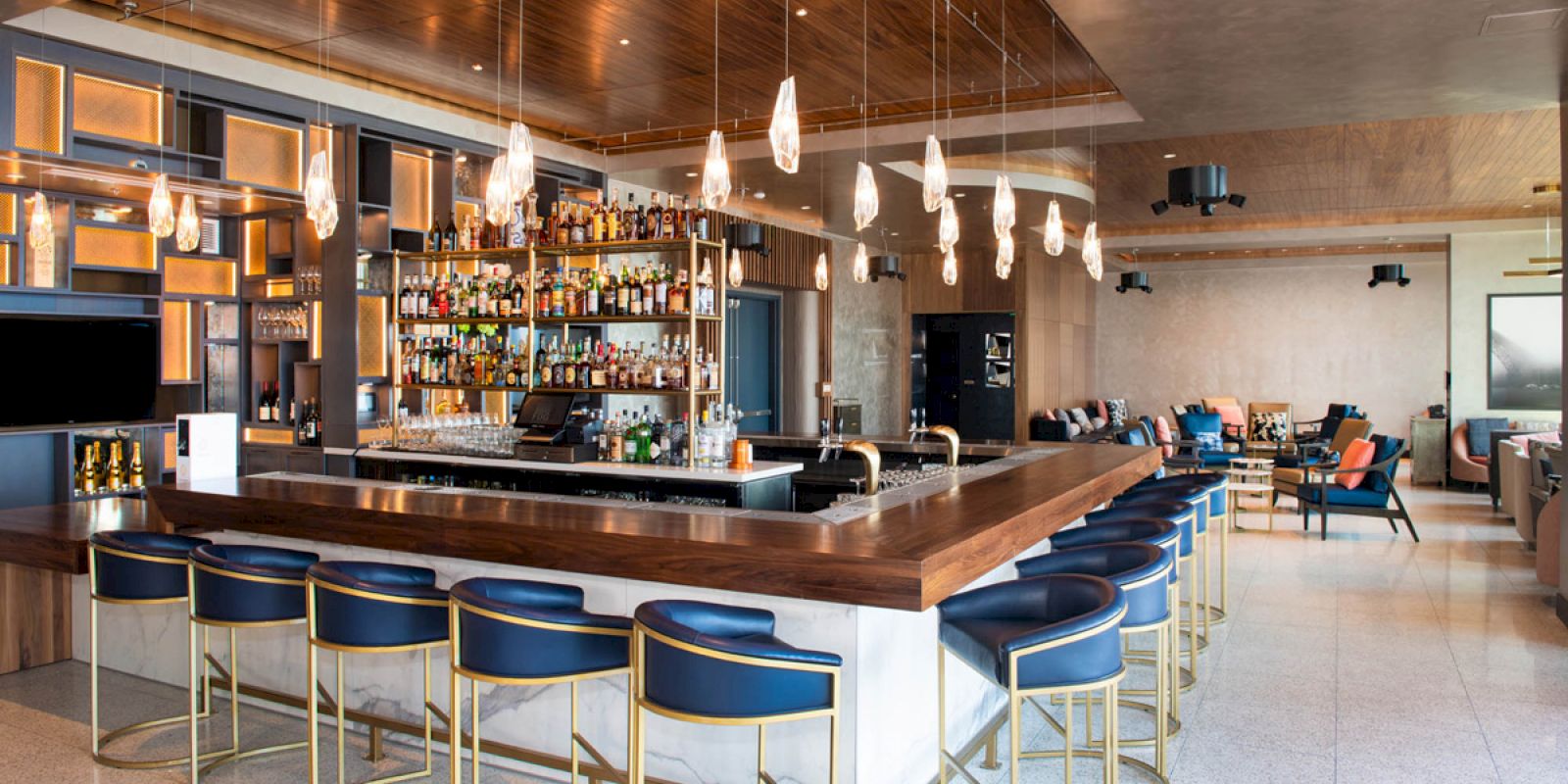 The image shows a modern bar with blue stools, hanging lights, and a well-stocked counter in an elegant, spacious setting.