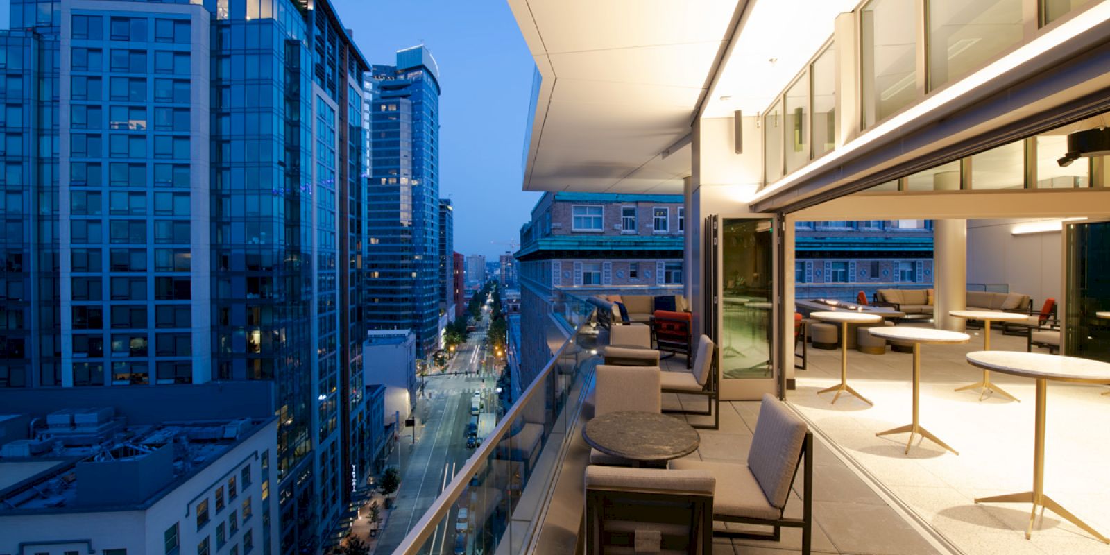 A modern rooftop patio with seating overlooks an urban cityscape at dusk, featuring tall buildings and a clear sky.