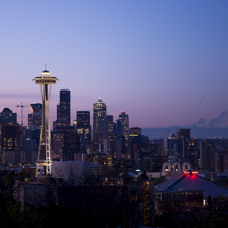 Seattle skyline at dusk with the Space Needle prominently visible, buildings, and Mount Rainier in the background.