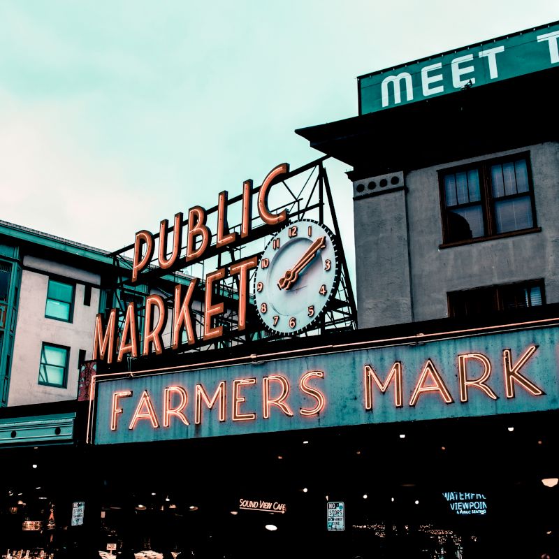 A neon sign for a public farmers market, with a clock, against the backdrop of urban buildings and a cloudy sky.
