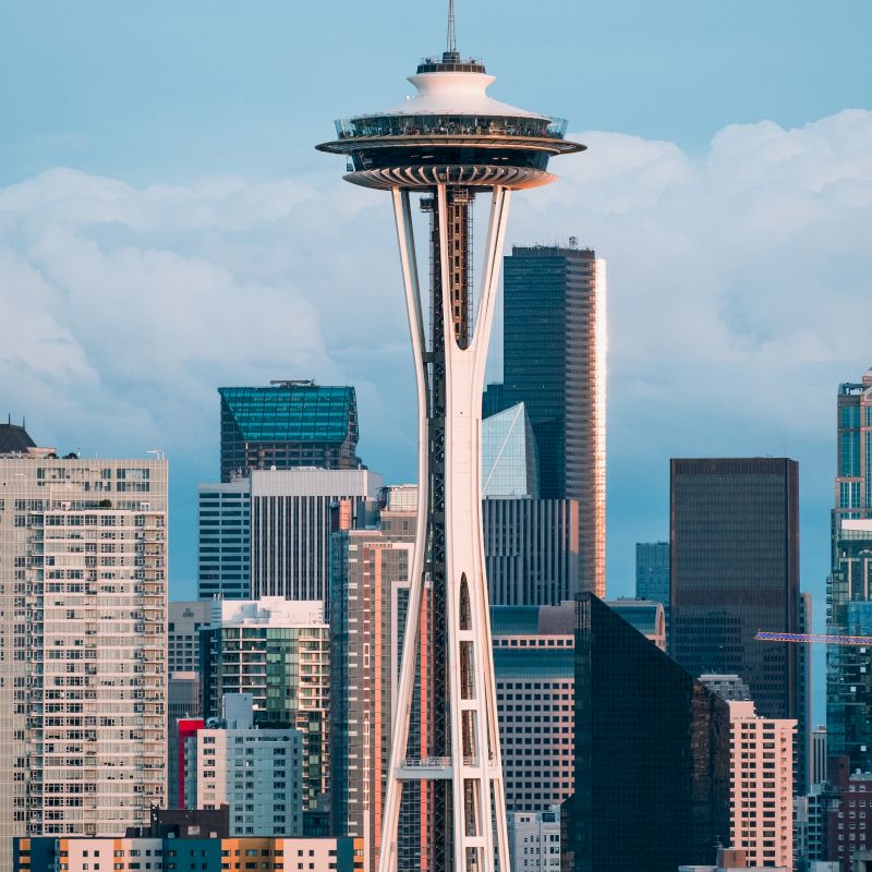 The image shows the Space Needle towering over the Seattle skyline, with various modern skyscrapers in the background under a cloudy sky.