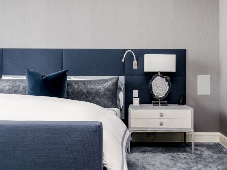 A modern bedroom features a bed with blue and white bedding, a nightstand with a lamp, and decorative elements on a carpeted floor.