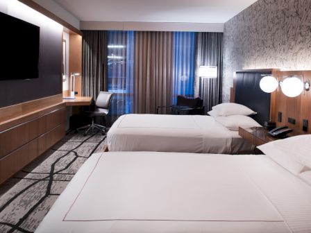 The image shows a modern hotel room with two beds, a large TV, a desk, and ambient lighting, creating a cozy and inviting atmosphere.