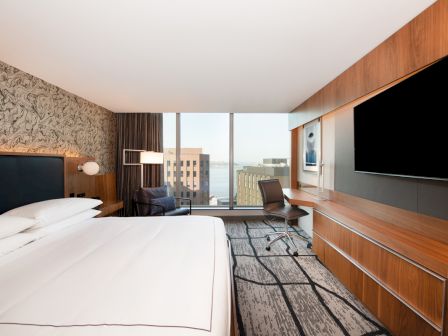 A modern hotel room with a large bed, wall-mounted TV, desk with chair, and window with city view, featuring stylish decor.