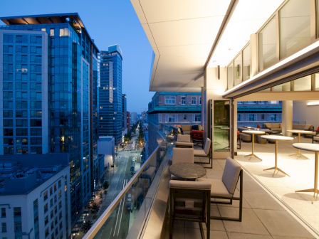 A modern balcony overlooking a cityscape at dusk, with tables and chairs for outdoor seating, adjacent to tall glass buildings.