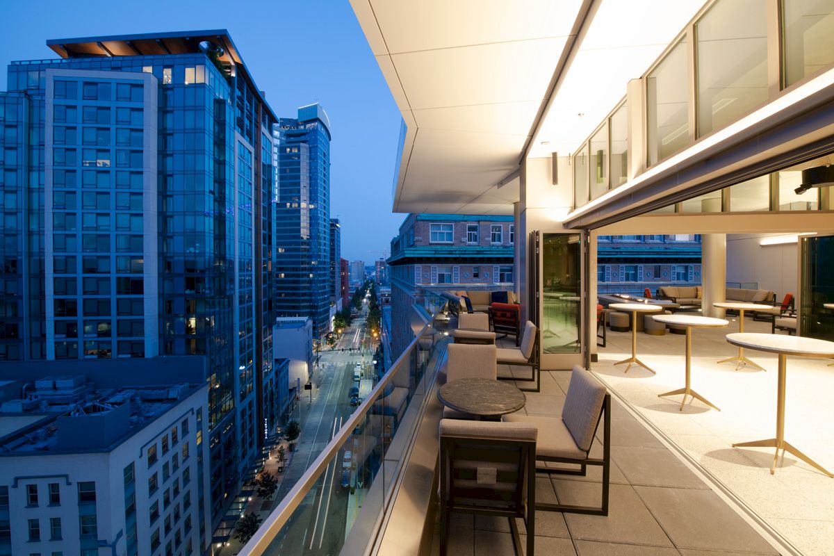 Cityscape view from a modern rooftop patio at dusk, featuring tables, chairs, and tall buildings lining the street below, creating a cozy ambiance.