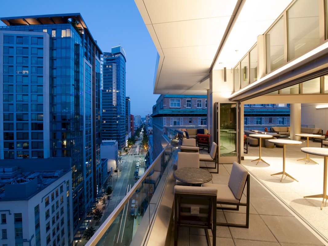 Cityscape view from a modern rooftop patio at dusk, featuring tables, chairs, and tall buildings lining the street below, creating a cozy ambiance.