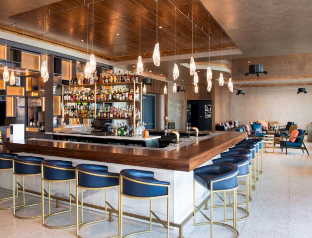 The image shows a stylish bar with blue stools, a wooden counter, and pendant lighting in a modern interior space.