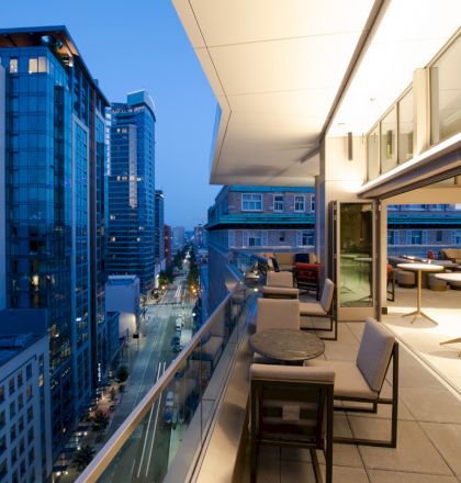 A modern rooftop patio with seating overlooks a city street lined with tall buildings, captured during the early evening.