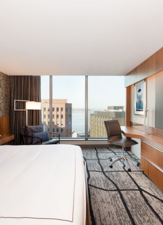 A modern hotel room features a bed, large TV, desk, and window with city view. The decor is contemporary with wood accents.