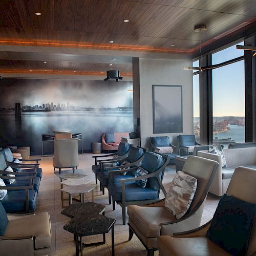 The image shows a modern lounge area with stylish seating, large windows, and a view of water and hills in the background.