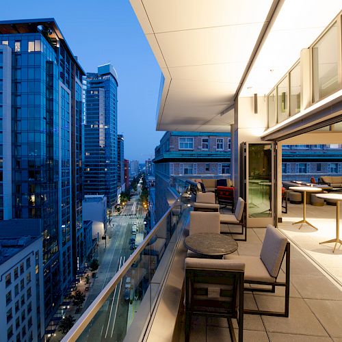 A modern urban balcony overlooks a cityscape with tall buildings and a street view at dusk, featuring seating areas and ambient lighting.