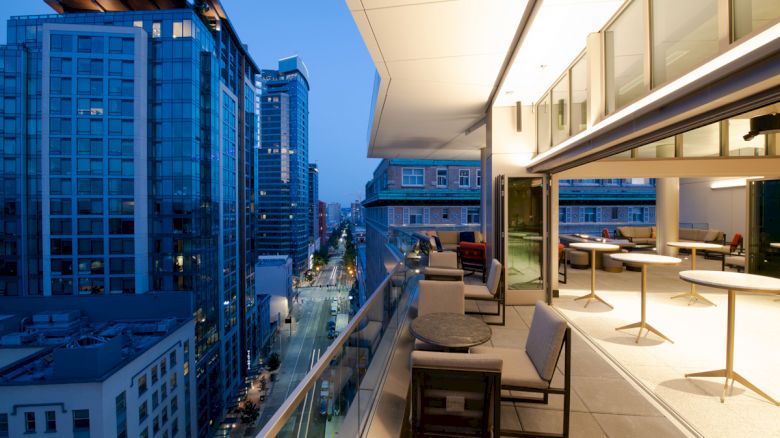 A modern rooftop terrace overlooks a cityscape with tall buildings, featuring outdoor seating and tables in the evening light.