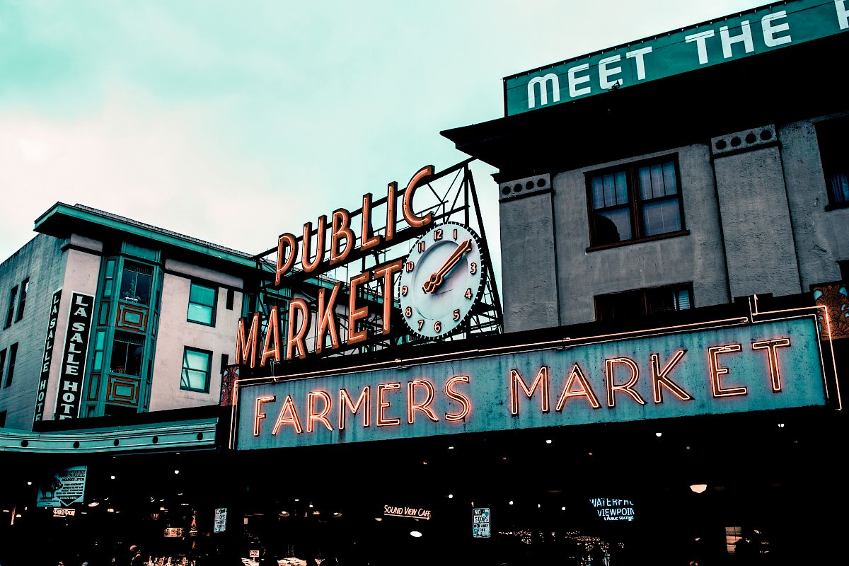 A public market sign with a clock and neon lights, advertising a farmers market in an urban setting.