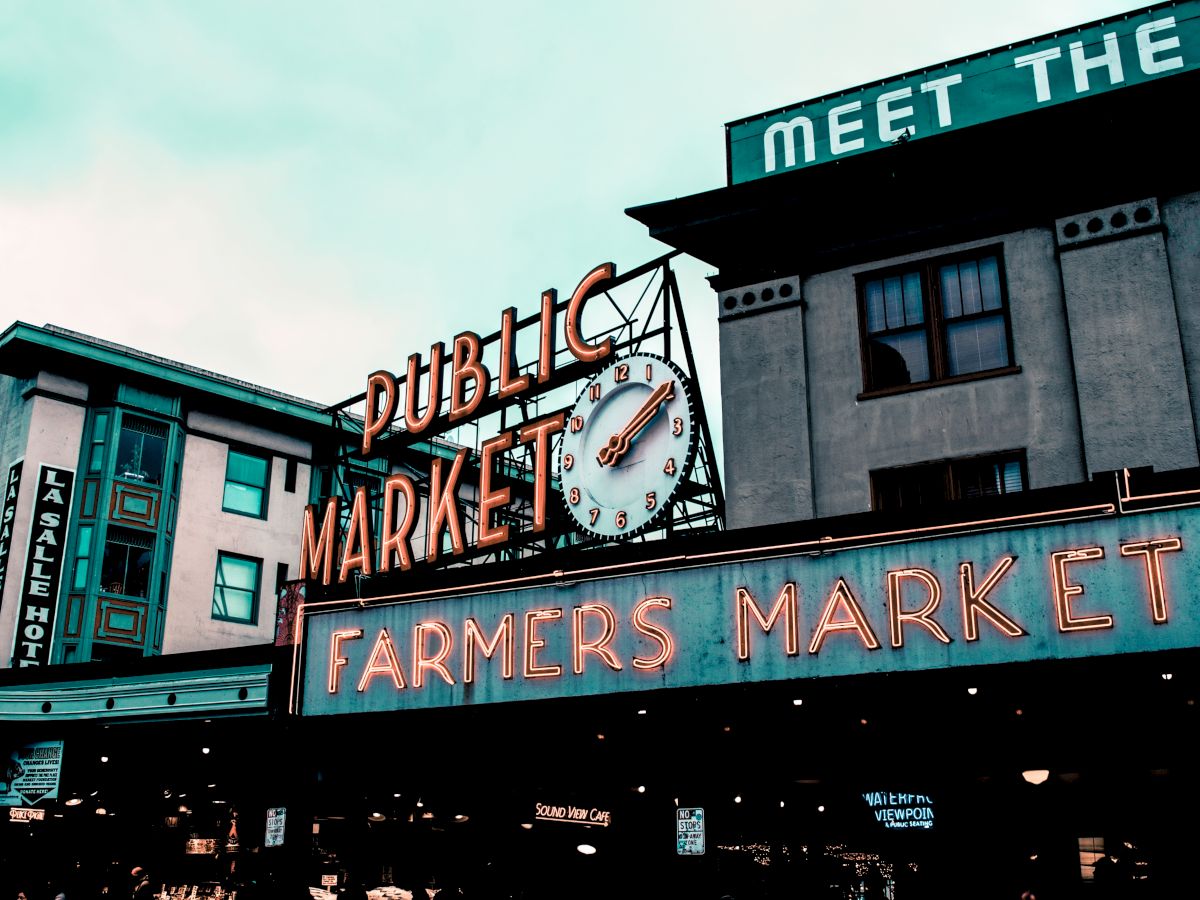 A public market sign with a clock and neon lights, advertising a farmers market in an urban setting.