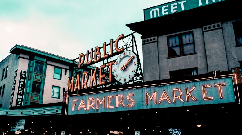 A public market sign with a clock and neon lights, advertising a farmers market in an urban setting.