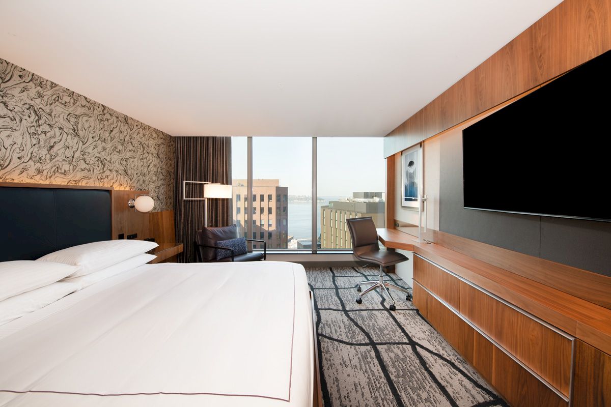 A modern hotel room featuring a large bed, wall-mounted TV, desk area, and large window with a cityscape view.