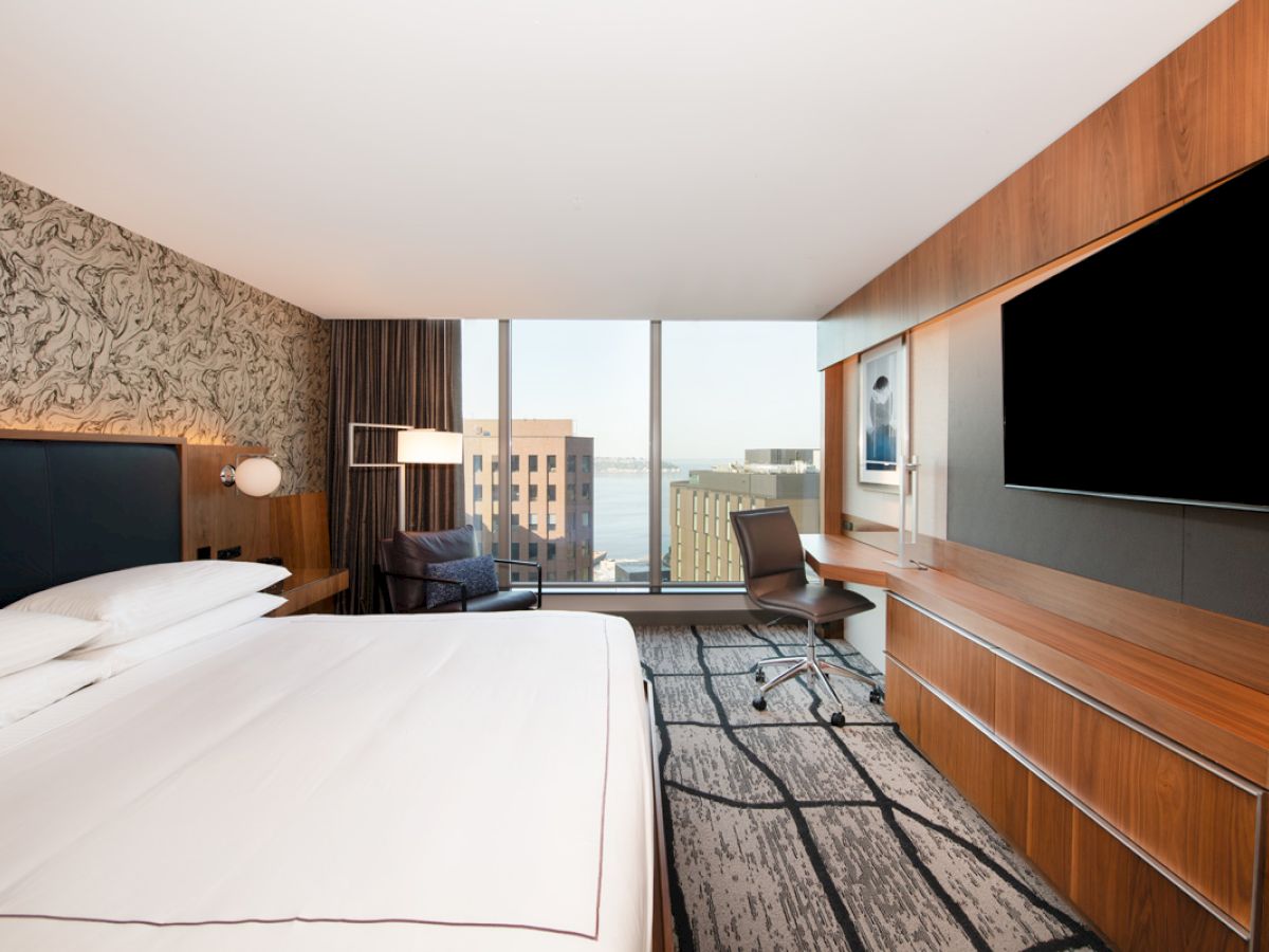 A modern hotel room featuring a large bed, wall-mounted TV, desk area, and large window with a cityscape view.