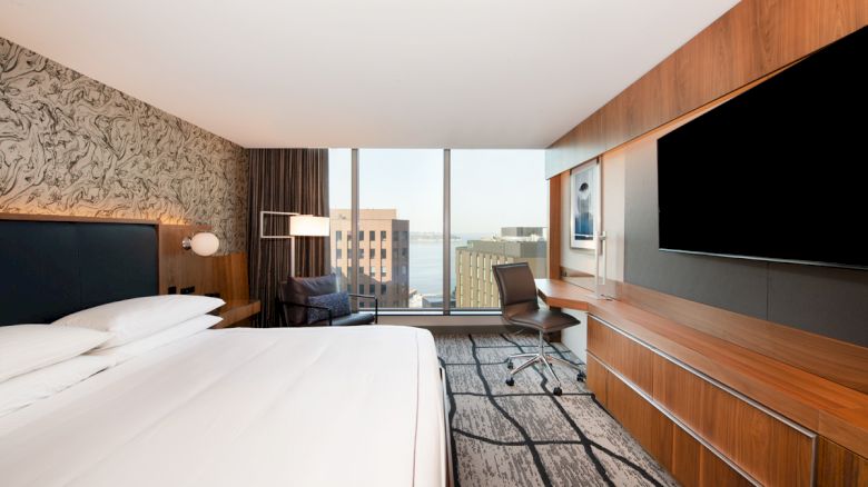 A modern hotel room featuring a large bed, wall-mounted TV, desk area, and large window with a cityscape view.