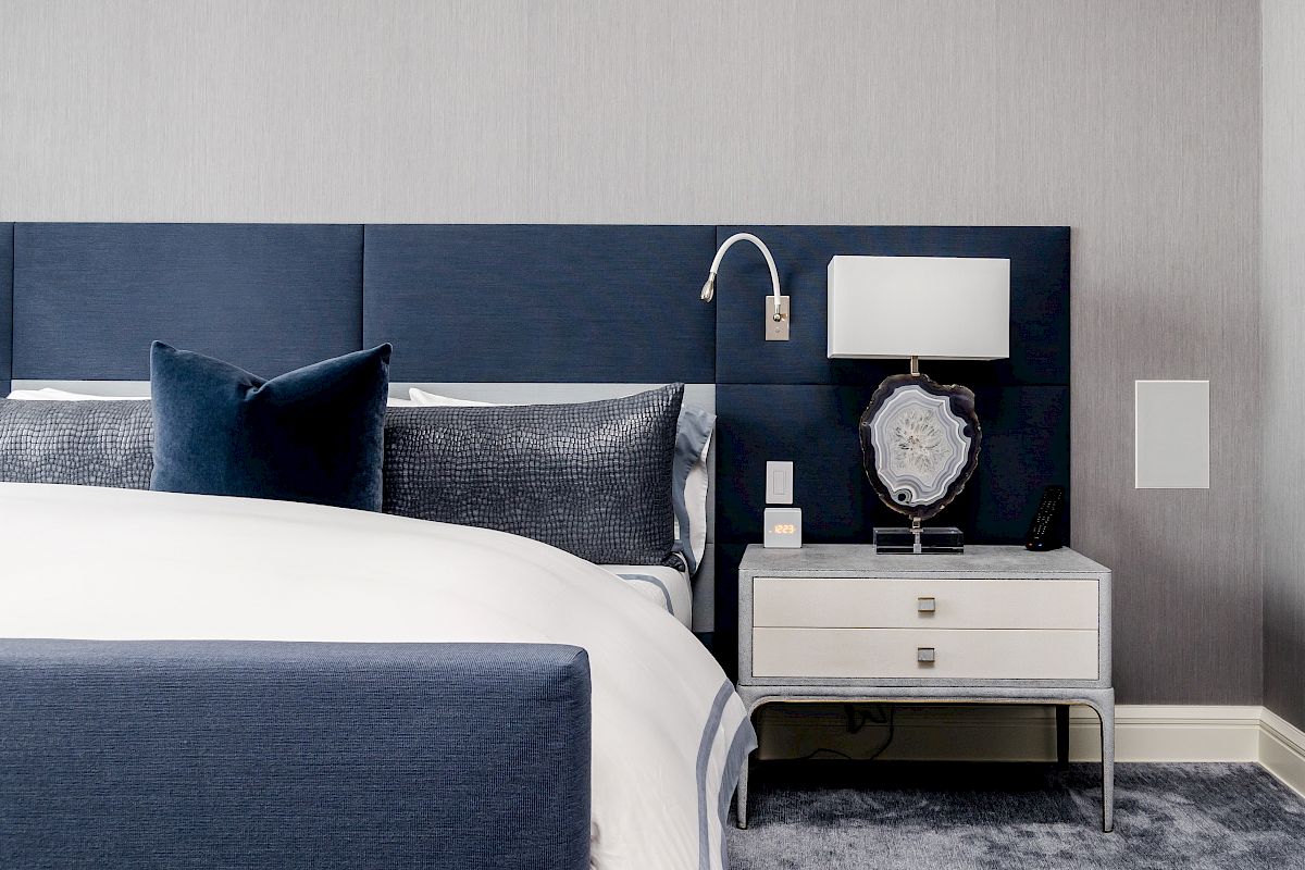 A modern bedroom with a navy headboard, white bedding, a bedside table, lamp, and an elegant decorative plate on display.