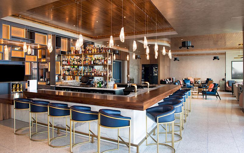 The image shows a modern bar with blue and gold stools, pendant lights, shelves stocked with bottles, and a lounge area in the background.