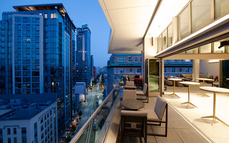 A modern rooftop terrace overlooking a cityscape at dusk, featuring seating areas with tables and chairs and illuminated high-rise buildings.