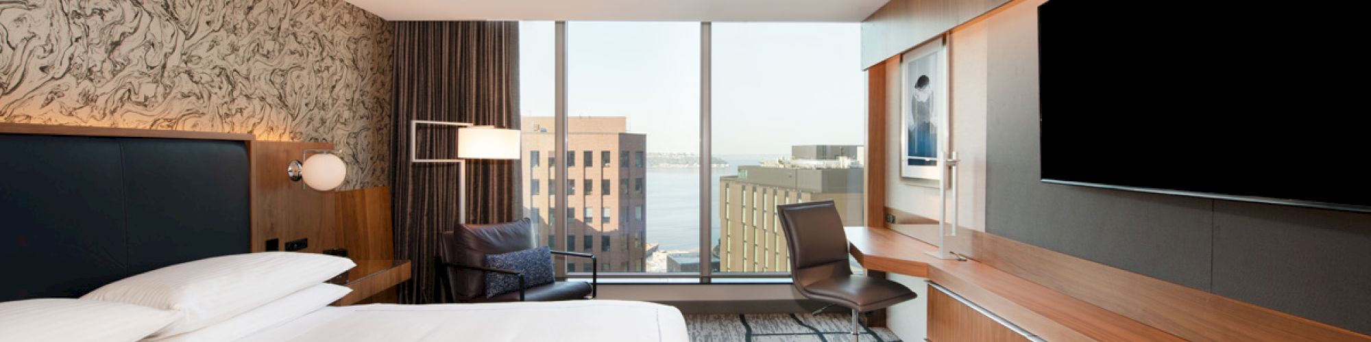 A modern hotel room with a large bed, desk, wall-mounted TV, and window overlooking a cityscape, accented with wood and neutral tones.